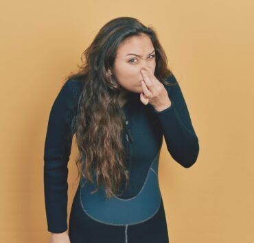 Young hispanic girl wearing neoprene scuba diving uniform smelling something stinky and disgusting, intolerable odor, holding breath with fingers on nose. bad smell