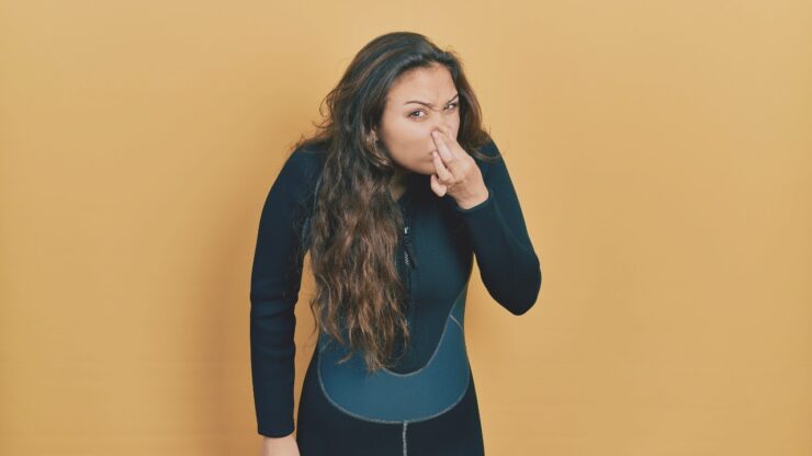 Young hispanic girl wearing neoprene scuba diving uniform smelling something stinky and disgusting, intolerable odor, holding breath with fingers on nose. bad smell