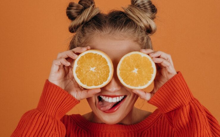 Mulher com suéter de tricô vermelho segurando duas fatias de laranja sobre os olhos