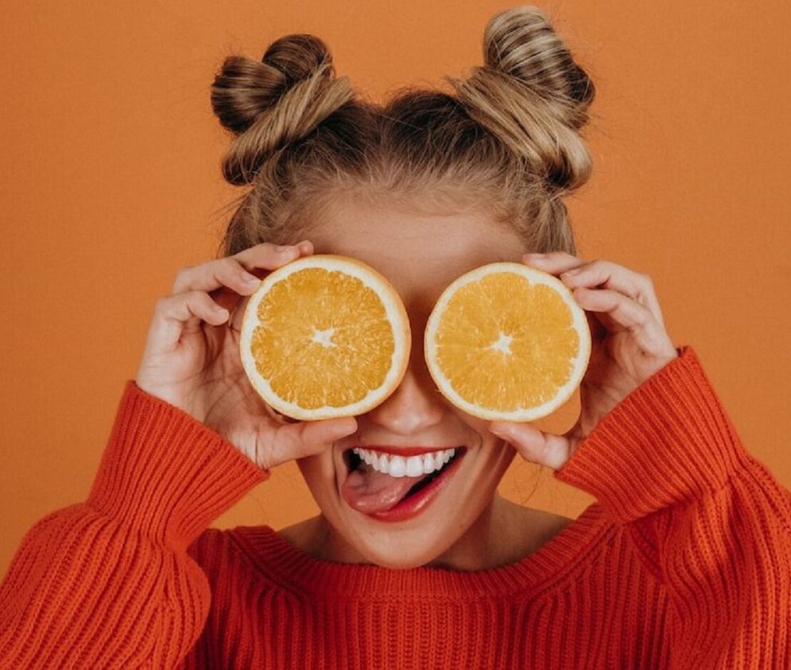 Mulher com suéter de tricô vermelho segurando duas fatias de laranja sobre os olhos