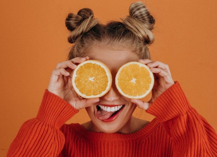 Mulher com suéter de tricô vermelho segurando duas fatias de laranja sobre os olhos