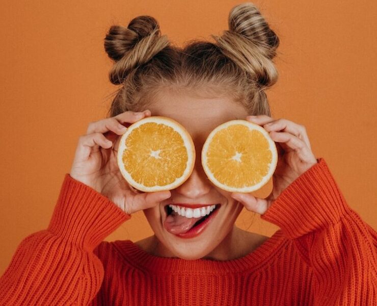 Mulher com suéter de tricô vermelho segurando duas fatias de laranja sobre os olhos