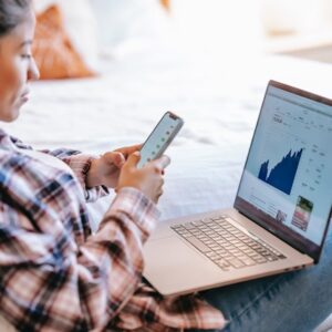 donna che guarda il cellulare sul letto con un laptop in fronte