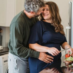 Cheerful pregnant wife in blue dress cooking sandwiches for breakfast while smiling and looking at husband embracing belly in modern kitchen